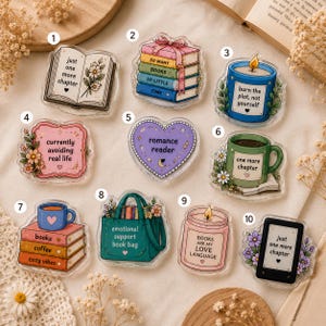 Könnte beinhalten: Zehn Acryl-Anstecknadeln mit Illustrationen und Text zum Thema Bücher. Die Designs umfassen ein offenes Buch, einen Bücherstapel, eine blaue Kerze, ein rosa Schild, ein lila Herz, eine grüne Tasse, einen Bücherstapel mit Tasse, eine Tragetasche, eine rosa Kerze und einen E-Reader.