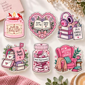 以下が含まれることがあります： ピンクを基調とした読書をテーマにしたイラストのデコレーション用アクリルステッカー6枚セット。テキスト入りのキャンドル、ハート型の本、本の上に置かれたヘッドフォン、ブックメールと書かれた郵便受け、薬瓶、アームチェアのデザインが含まれています。