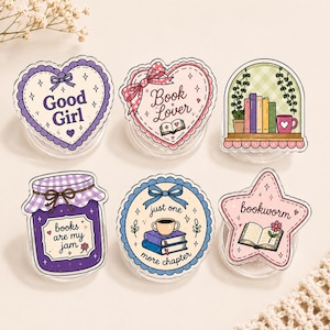 Puede incluir: Seis soportes decorativos para teléfono con ilustraciones coloridas. Los diseños incluyen un corazón morado con 'Good Girl', un corazón rosa con 'Book Lover', una estantería, un tarro de mermelada morado con 'books are my jam', un círculo azul con 'just one more chapter' y una estrella rosa con 'bookworm'.