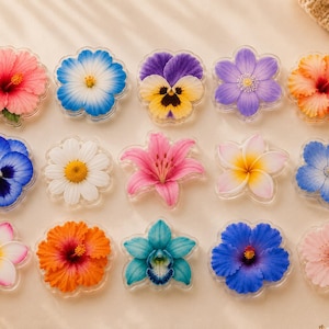 May include: A collection of fifteen individual flower-shaped acrylic or resin decorative pieces arranged in three rows. The assortment features various floral designs including hibiscus, pansies, daisies, lilies, and plumeria in vibrant colors.