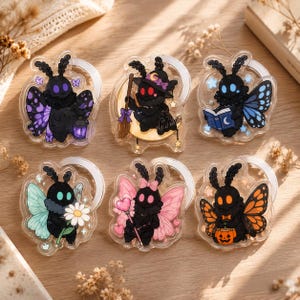 May include: Six clear acrylic charms featuring black moth characters with various accessories. One holds a purple lantern, one sits on a crescent moon with a broom, one reads a book, one holds a daisy, one holds a heart wand, and one holds a pumpkin.