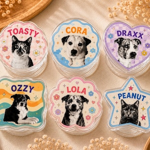 May include: Six clear acrylic phone grips featuring custom pet portraits. Each piece displays a unique shape, including circles, a heart, a star, and a paw print. The designs include the names Toasty, Cora, Draxx, Ozzy, Lola, and Peanut.