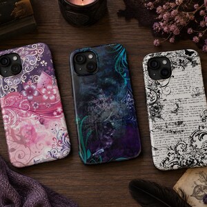 May include: Three smartphone cases featuring distinct designs: one with pink and purple floral swirls, one with a dark blue and teal abstract pattern, and one with black vintage-style script text and ornate flourishes on a white background.