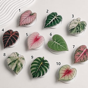 May include: Ten circular phone grips featuring various leaf designs labeled 1 through 10. The designs include pink, green, and white variegated foliage patterns shaped like hearts or ovals, each with a glossy finish and a white base.