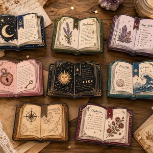 May include: A collection of eight rectangular enamel pins shaped like open books, each featuring unique illustrations including a crescent moon, lavender, crystals, a heart lock, a sun, ocean waves, a compass, and roses with detailed text.