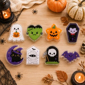 Op de afbeelding: Een verzameling van acht acrylspeldjes met Halloween-figuren, waaronder een witte geest met een pompoen, een groen Frankenstein-hart, een oranje ster-pompoen, een vampier in een doodskist, een zwarte kat op een maan, een mummie, een Magere Hein en een paarse vleermuis.