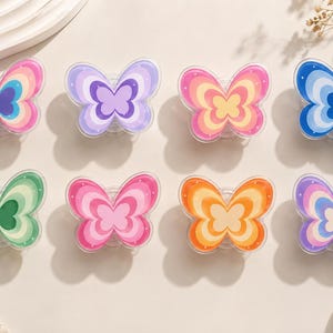 May include: A collection of eight butterfly-shaped phone grips arranged in two rows. Each grip features a distinct color palette with concentric layered patterns, including shades of pink, purple, yellow, blue, green, and orange with white dots.