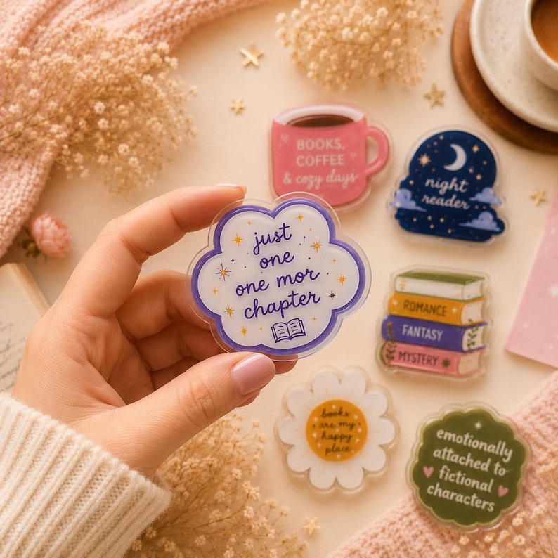 May include: A collection of acrylic pins featuring various designs including a pink mug with text, a night sky scene, a stack of books, a daisy flower, a green scalloped shape with text, and a white scalloped pin reading just one one mor chapter.