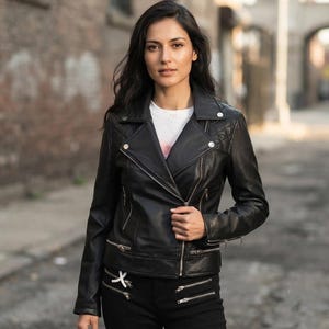 May include: A black leather motorcycle jacket featuring an asymmetrical silver zipper, quilted shoulder panels, and multiple zippered pockets. The jacket is worn over a white t-shirt, paired with black pants that have horizontal zippered accents.