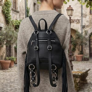 May include: A black backpack with a textured surface, featuring long fringe detailing on the sides, silver-toned metal hardware, a zipped pocket on the back, and adjustable shoulder straps adorned with multiple circular metal rings.