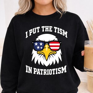May include: A black long-sleeve sweatshirt featuring a graphic of a bald eagle wearing sunglasses with an American flag pattern. The text I PUT THE TISM IN PATRIOTISM is printed in white capital letters above and below the eagle illustration.