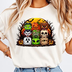 May include: A cream-colored t-shirt featuring a graphic print of eight block-shaped Halloween characters, including a green monster, orange pumpkin, brown werewolf, vampire, zombie, skull, ghost, and alien, set against a large yellow moon.