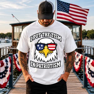 May include: A white short-sleeved t-shirt featuring a graphic of a bald eagle wearing sunglasses with American flag patterns. The text above the eagle reads I PUT THE TISM and the text below reads IN PATRIOTISM in bold black capital letters.