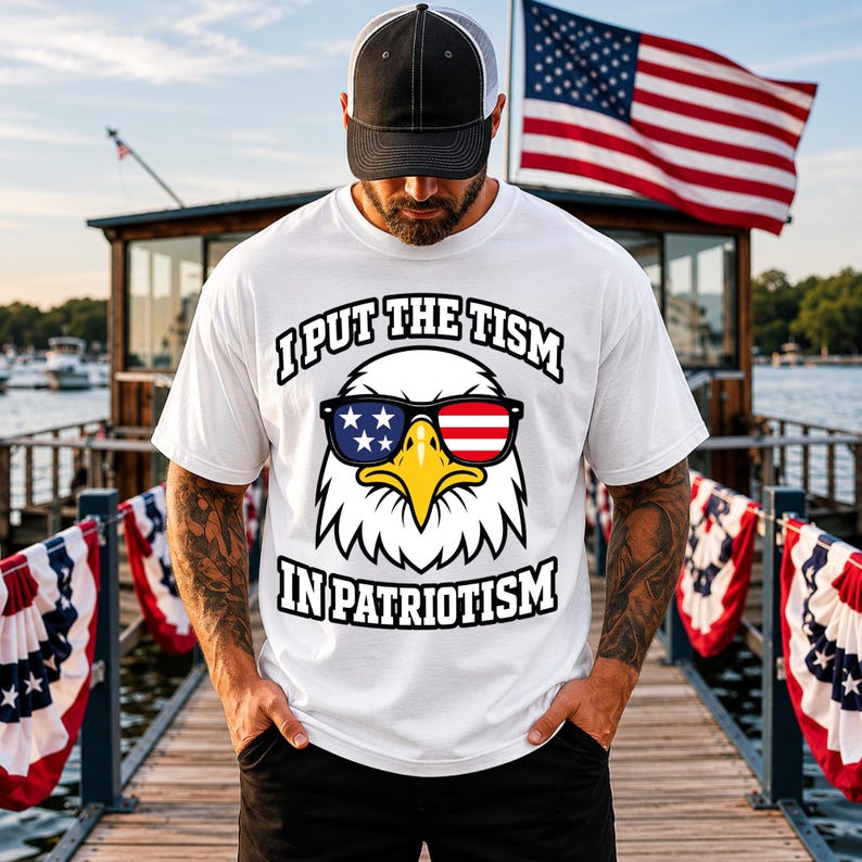 May include: A white short-sleeved t-shirt featuring a graphic of a bald eagle wearing sunglasses with American flag patterns. The text above the eagle reads I PUT THE TISM and the text below reads IN PATRIOTISM in bold black capital letters.