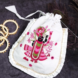 May include: A white drawstring pouch featuring an embroidered figure in pink, green, and black thread. The bag is accented with a ruffled white trim and gold sequins along the edges. The text KAYATARAA is printed in white above the pouch.
