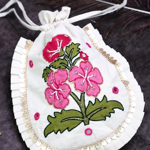 May include: A white drawstring pouch featuring the text KAYATARAA, decorated with embroidered pink flowers and green leaves. The edges are trimmed with pleated white fabric and gold sequins, with small pink circular accents scattered throughout.