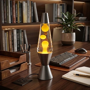 Op de afbeelding: Een lavalamp met een zilverkleurige metalen voet en dop, voorzien van een glazen reservoir gevuld met vloeistof en zwevende gele wasbollen. De lamp heeft een conische vorm, een stroomsnoer en verspreidt een warme gloed vanuit de lichtbron.