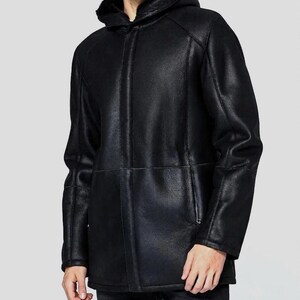 Könnte beinhalten: Eine schwarze Kapuzenjacke mit glatter, lederartiger Außenfläche und sichtbarem dunklem Innenfutter. Die Jacke verfügt über eine mittige vertikale Naht, lange Ärmel und Seitentaschen, was eine strukturierte Silhouette mit Kapuze ergibt.