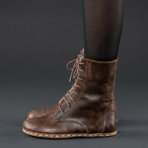 Könnte beinhalten: Ein dunkelbrauner Schnürstiefel aus Leder mit abgerundeter Zehenpartie und sichtbaren hellen Nähten entlang der Sohle. Der Stiefel hat ein hohes Design mit dünnen braunen Schnürsenkeln, die durch Metallösen an der Vorderseite verlaufen.