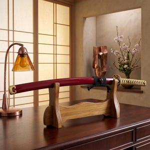 May include: A katana sword with a glossy red scabbard and a gold-colored hilt, resting horizontally on a light brown wooden display stand. The sword features a black cord wrap near the hilt and ornate gold-toned metal accents at both ends.