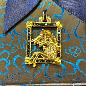 Könnte beinhalten: Ein goldener Anhänger mit zwei Pferdeköpfen in einem rechteckigen Rahmen. Der Rahmen ist mit dunkelblauen, quadratisch geschliffenen Steinen besetzt. Die Pferde sind mit klaren Pavé-Steinen und kleinen roten Steinen als Augen verziert.