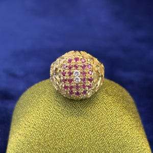 Puede incluir: Un anillo de oro con una superficie redondeada y texturizada que presenta un grupo ovalado de pequeños rubíes de color rosa vibrante dispuestos en cuadrícula, con dos diamantes blancos redondos y claros engastados verticalmente.