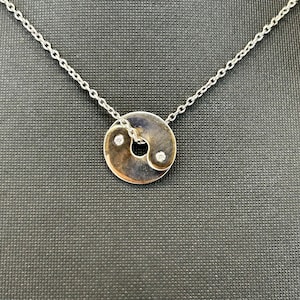 May include: A silver-toned necklace featuring a circular pendant with a yin-yang design. The pendant is divided into two contrasting sections, each containing a small, clear, round-cut gemstone. The pendant hangs from a delicate silver-toned chain.
