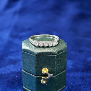 May include: A silver-toned metal ring featuring five small, clear, round-cut stones set in a row along the top band. The ring is positioned on top of a dark green, hexagonal-shaped jewelry box with a gold-toned metal latch on the front side.