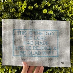 May include: A rectangular needlepoint canvas featuring a light blue stitched border and the text THIS IS THE DAY THE LORD HAS MADE LET US REJOICE & BE GLAD IN IT! in light blue lettering. The initials FM are stitched in the bottom right corner.