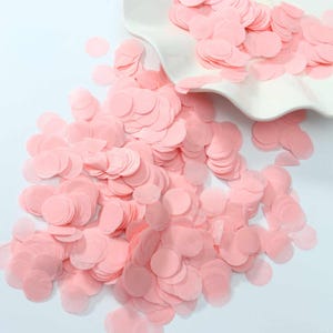 May include: A large collection of small, round, light pink paper confetti pieces scattered across a surface. The thin, circular paper discs are uniform in shape and size, creating a dense pile of soft pink color that appears light and airy.