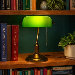 May include: A green-shaded desk lamp with a brass base and stem sits on a wooden shelf. The lamp is positioned next to a small potted plant and a row of books, including "RALPH LAUREN" and "THE WATCH BOOK". The background is a dark green wall.