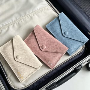 Op de afbeelding: Drie portemonnees in envelopstijl met een generfde textuur, voorzien van een drukknoopsluiting op de flap. De portemonnees zijn uitgevoerd in crème, oudroze en lichtblauw. De crème en roze portemonnees zijn voorzien van kleine initialen.