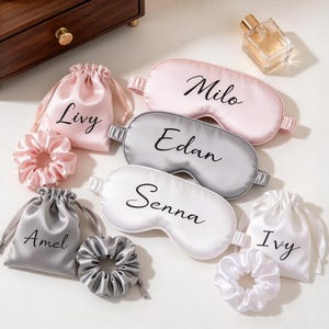 May include: A collection of three satin sleep masks in pink, gray, and white, each featuring a name in black script: Milo, Edan, and Senna. Included are three matching drawstring pouches labeled Livy, Amel, and Ivy, along with three matching scrunchies.