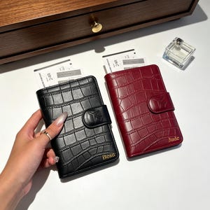 May include: Two passport holders, one black and one deep red, featuring a crocodile-embossed texture and a snap closure with an airplane icon. The black holder is personalized with the name Beau, and the red holder with the name Jude in gold.