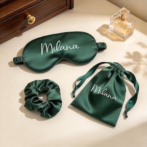 May include: A dark green satin sleep mask, a matching dark green satin scrunchie, and a dark green satin drawstring pouch. The sleep mask and the pouch both feature the name Milana printed in white cursive script lettering on the front surface.
