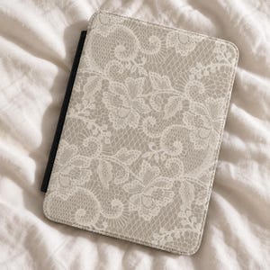Puede incluir: Una funda rectangular para tableta con un patrón de encaje blanco impreso sobre un fondo beige. El estuche tiene un lomo negro en el lado izquierdo y esquinas redondeadas, ofreciendo una cubierta protectora para un dispositivo.