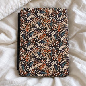 Puede incluir: Una funda rectangular para lector electrónico con un patrón repetitivo de alas de mariposa en tonos negro, crema, naranja y azul verdoso. La funda tiene un lomo negro y una superficie texturizada con un diseño denso de motivos de alas.