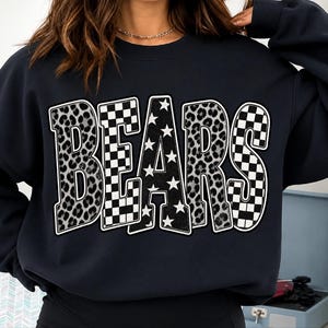May include: A dark navy blue crewneck sweatshirt featuring the word BEARS printed across the chest. The letters are filled with a mix of gray and black leopard print, black and white checkered patterns, and a black background with white stars.