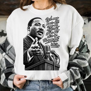 Może przedstawiać: Biała bluza z okrągłym dekoltem z czarno-białą grafiką przedstawiającą Dr. Martina Luthera Kinga Jr. przy mównicy z mikrofonem. Tekst brzmi: We may have all come on different ships but we're in the same boat. Dr. Martin Luther King Jr.