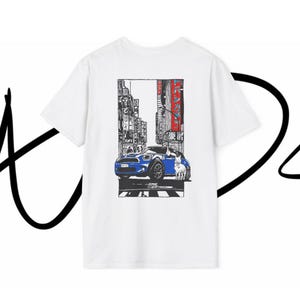 Puede incluir: Una camiseta blanca de manga corta con un gráfico rectangular en la espalda. El estampado muestra un coche azul aparcado en una calle urbana con edificios altos, texto en japonés en los carteles y un perro blanco junto al vehículo.