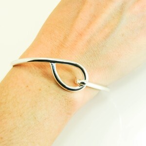 May include: A silver bangle bracelet with a teardrop-shaped design. The bracelet is open and has a simple, elegant style.