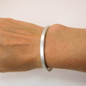May include: A silver bracelet with a brushed finish. The bracelet is open and has a simple, minimalist design.