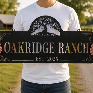 May include: A rectangular black metal sign featuring a laser-cut tree silhouette at the top center. The text OAKRIDGE RANCH is cut out in the center, with EST. 2025 below it. Decorative scrollwork accents the top left and right corners of the sign.