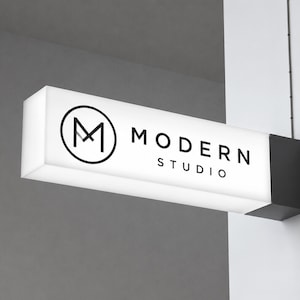 May include: A rectangular, illuminated wall-mounted sign with a white face and black side bracket. The sign features a black circular logo containing the letter M, followed by the black text MODERN STUDIO in a clean, sans-serif typeface.