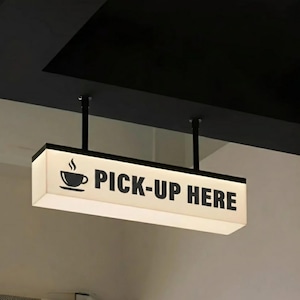 May include: A rectangular, illuminated hanging sign suspended by two black rods from a ceiling. The sign features a white face with black text that reads PICK-UP HERE, accompanied by a black graphic icon of a steaming coffee cup on a saucer.