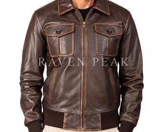 Könnte beinhalten: Eine dunkelbraune Lederjacke mit durchgehendem Frontreißverschluss, klassischem Kragen und zwei Brusttaschen mit Knopfklappen. Das Kleidungsstück hat gerippte Bündchen und einen gerippten Saum, mit dem Schriftzug RAVEN PEAK auf der Brust.