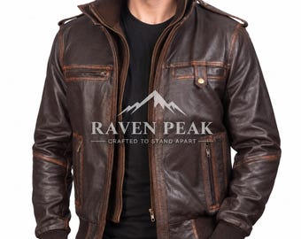 Könnte beinhalten: Eine dunkelbraune Lederjacke mit hohem Kragen, gerippten Bündchen und doppeltem Reißverschluss. Die Jacke verfügt über mehrere Reißverschlusstaschen an Brust und Seiten, Schulterklappen und den Text RAVEN PEAK CRAFTED TO STAND APART.