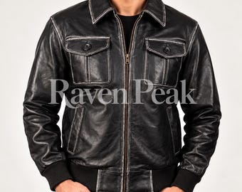 Könnte beinhalten: Eine schwarze Lederjacke mit Used-Look, ausgestattet mit einem durchgehenden Frontreißverschluss, einem spitzen Kragen und zwei Brusttaschen mit Knopfklappen. Die Jacke hat schwarze Rippbündchen an Ärmeln und Saum, mit dem Text Raven Peak.