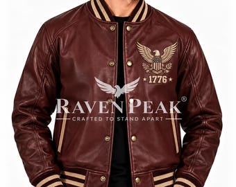Könnte beinhalten: Eine dunkelmarone Leder-Collegejacke mit geripptem Kragen und Bündchen mit beigen Streifen. Auf der Brust befindet sich ein goldenes Adleremblem über dem Text 1776. Die Jacke hat Metalldruckknöpfe und den Raven Peak Logo-Schriftzug.