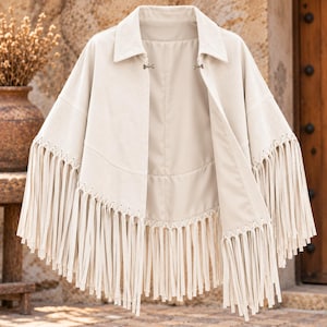 Puede incluir: Un poncho de color crema con cuello en punta y cierre de gancho en la parte delantera. La prenda presenta largos flecos verticales a lo largo del dobladillo y en la sección media, creando una apariencia texturizada y superpuesta.